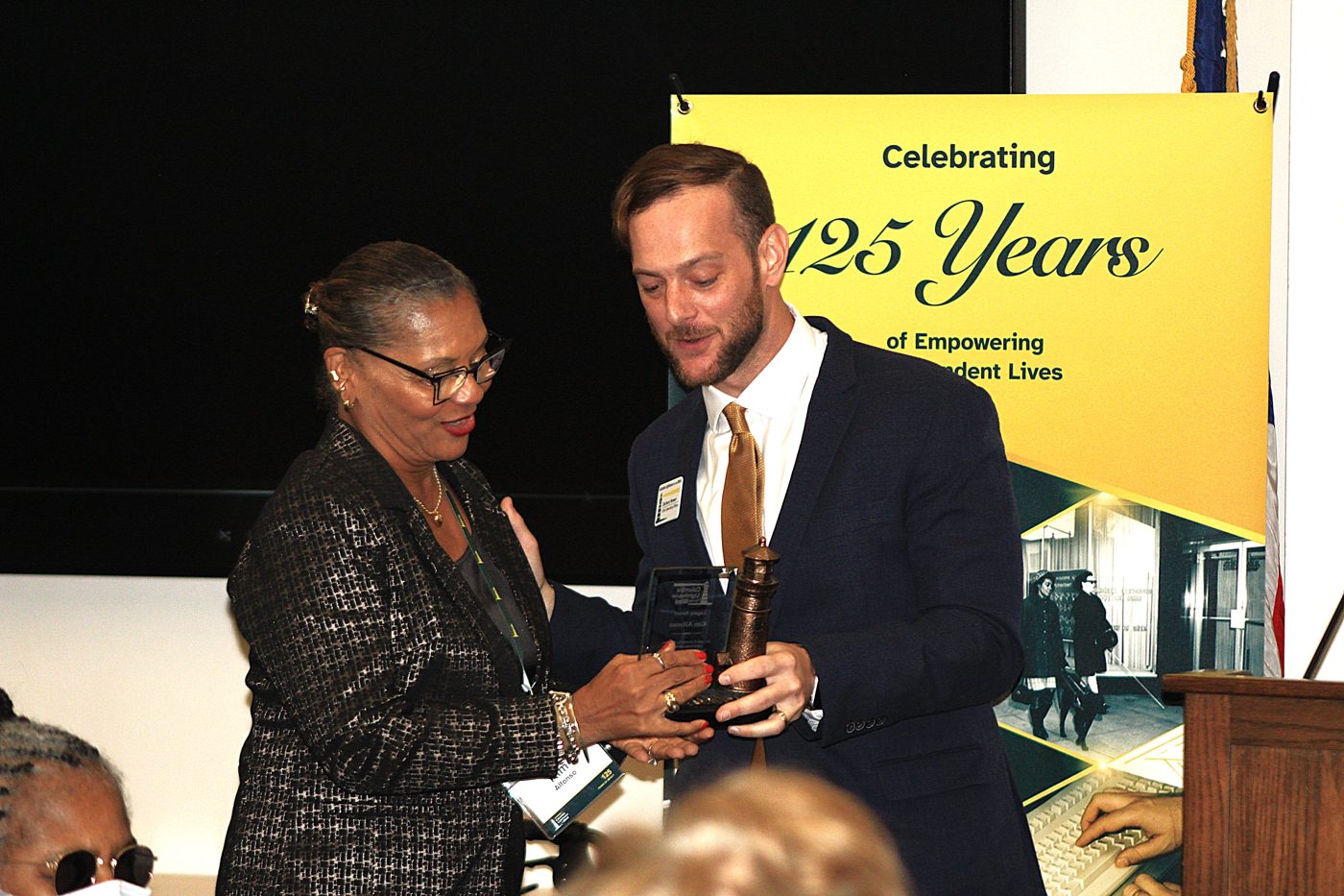 Kim Alfonso receives her bronze lighthouse Legacy Award from COO Zachary Brewer in front of the Celebrating 125 Years banner.