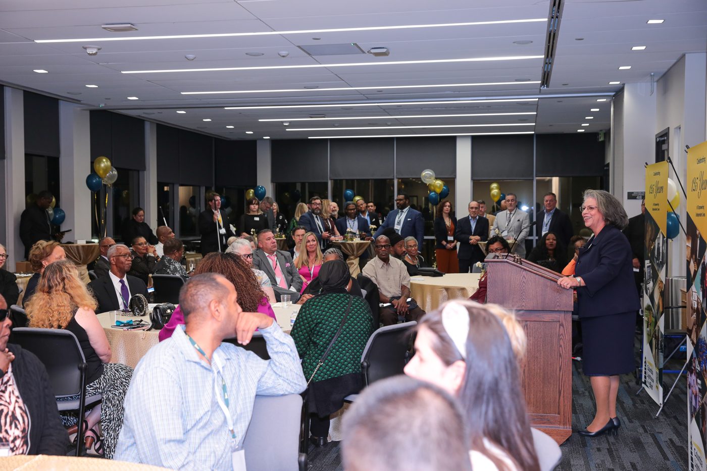 Soraya speaks from the podium to a full room of guests at round tables, with balloons and a 125 Years banner.