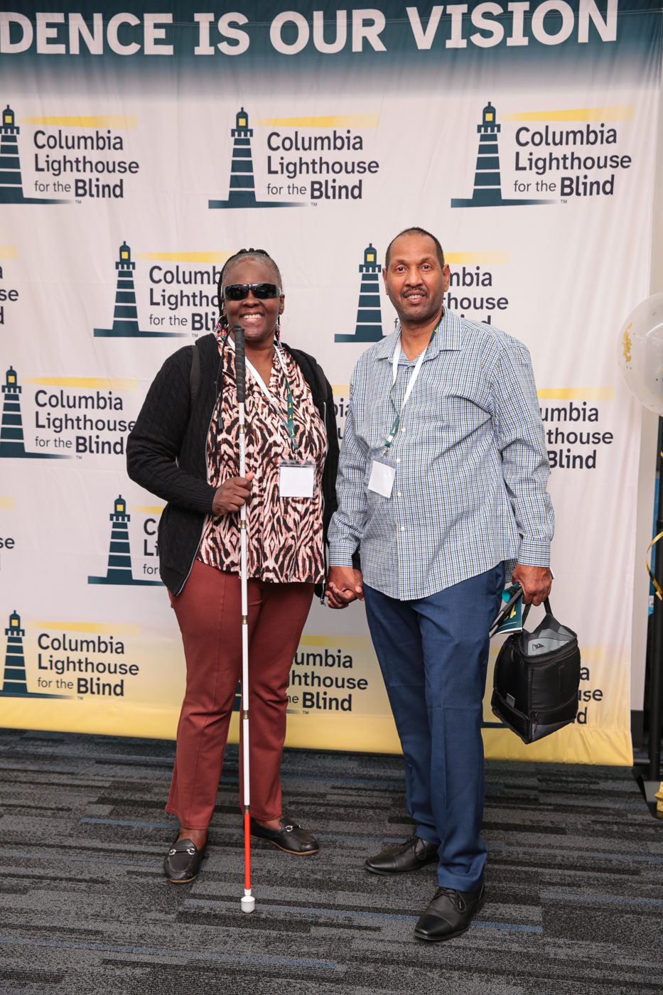 A smiling guest with a white cane and her companion stand together in front of the Independence Is Our Vision wall of Columbia Lighthouse for the Blind logos.