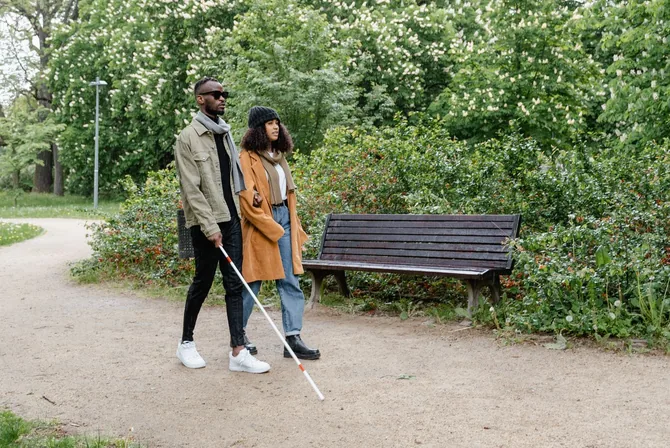 A Black man who is blind walks confidently along a tree-lined park path using a long white cane, accompanied by a woman beside him on a bright spring day.