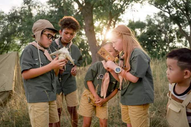 A diverse group of children in summer camp uniforms examine an animal skull and nature-study cards together in a sunlit grassy field during an outdoor camp activity.