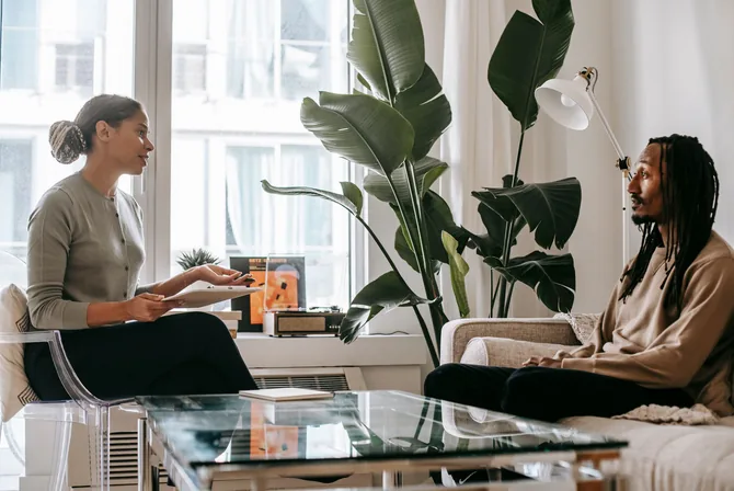 A Black case manager holding a document sits and talks with a Black client seated across from her in a bright, plant-filled living room during a one-on-one support meeting.