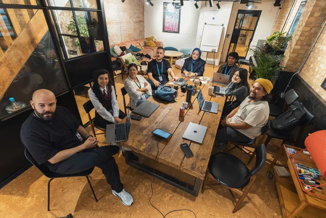 A diverse team of young professionals works on laptops around a large reclaimed-wood table in a modern brick-walled workspace.