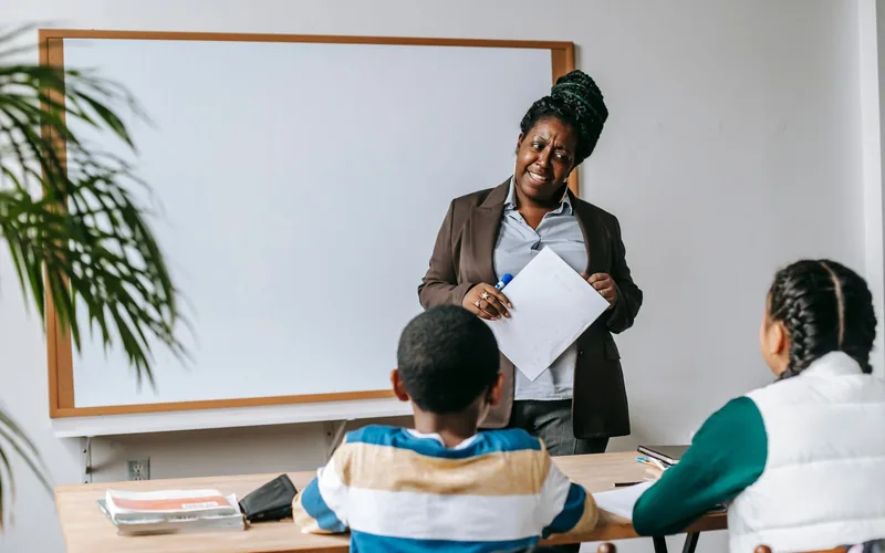 A Black teacher stands at a whiteboard and works with two students seated at a table in a bright classroom during a skills workshop.
