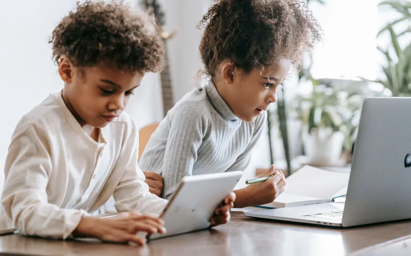 Two Black children lean over a table together, one using a tablet and the other a laptop, exploring technology side by side.