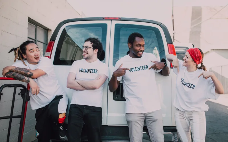 Four volunteers of different backgrounds wearing shirts that read Volunteer stand together laughing beside a van.