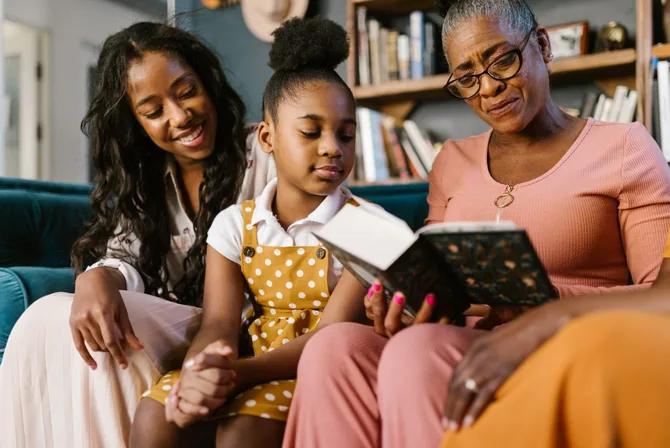 Three generations of a Black family, a young mother, a girl, and her grandmother, sit close together reading a book at home in front of a full bookshelf.