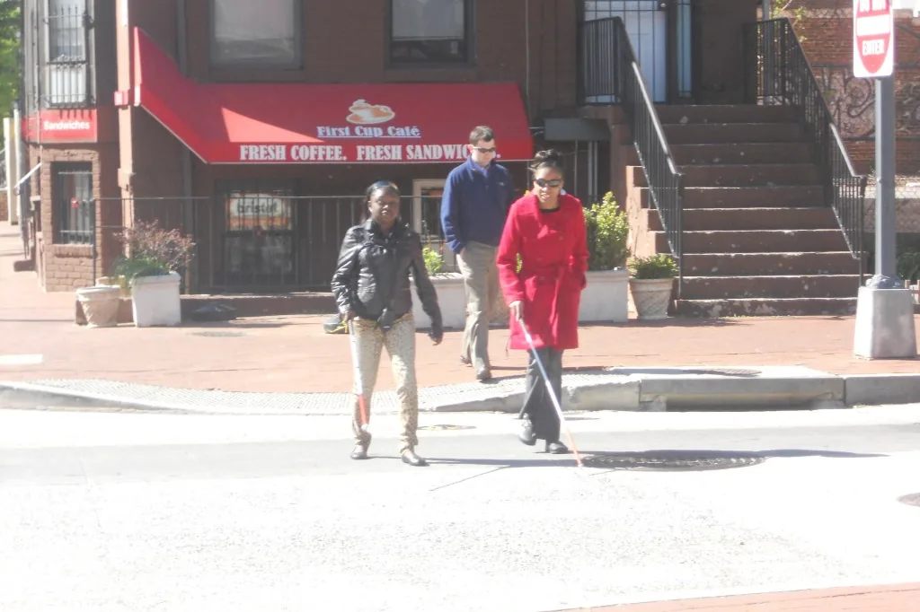 A woman using a long white cane crosses a neighborhood street near a corner cafe, traveling independently, with two other people walking nearby.