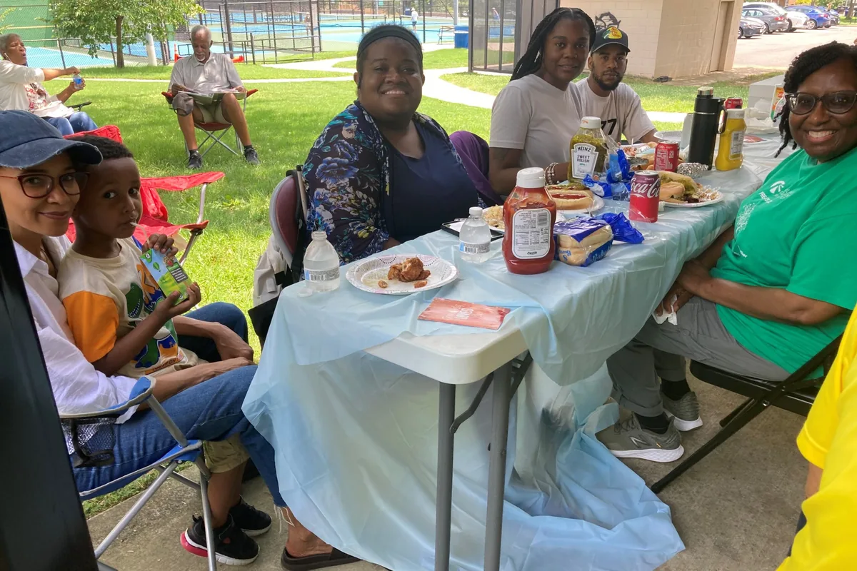 A CLB staff member in a green shirt sits with families and community members, including a young child, at outdoor picnic tables during a Mobile Eye Care community outreach event.