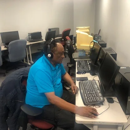 Solomon, a man in a bright blue polo shirt wearing headphones, working at a desktop computer in CLB's Silver Spring training lab.