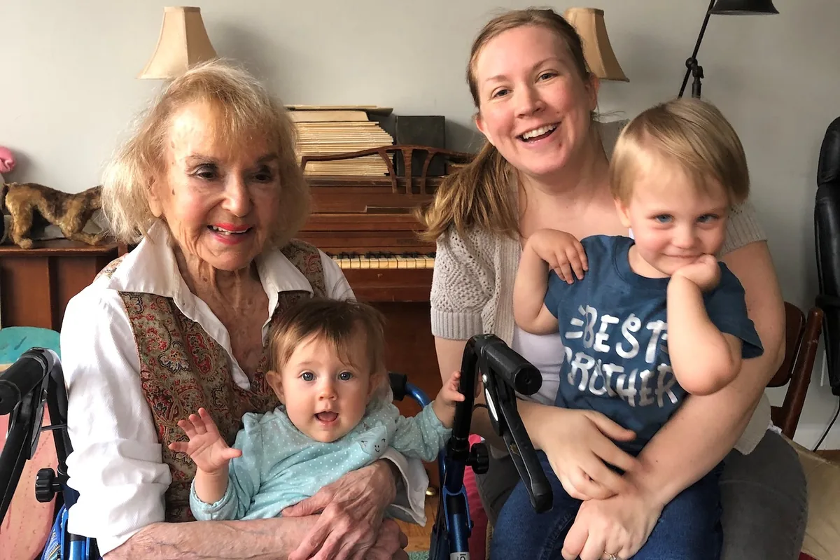 A volunteer and a young family visit an older woman who uses a walker at home, gathered together and smiling in her living room with two young children.