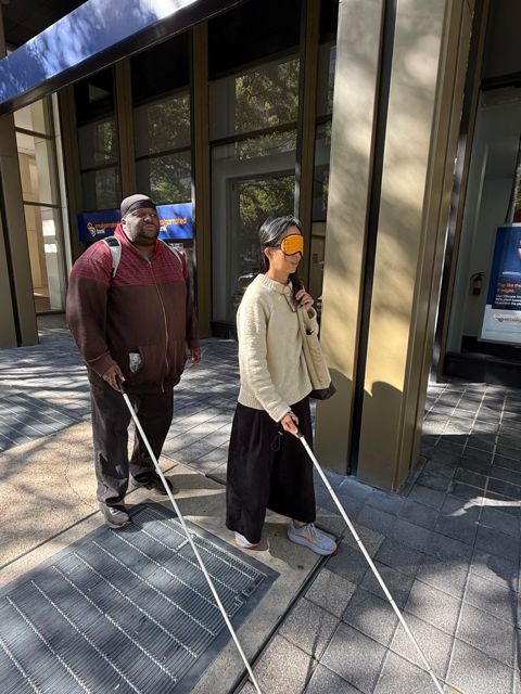 Alisa Tang, blindfolded and holding a white cane, walks a downtown D.C. sidewalk beside Antonio Williams, a CLB orientation and mobility specialist who is blind.