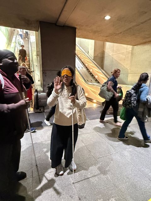 Alisa, still blindfolded, smiles and waves at the bottom of the Metro escalator next to Antonio.